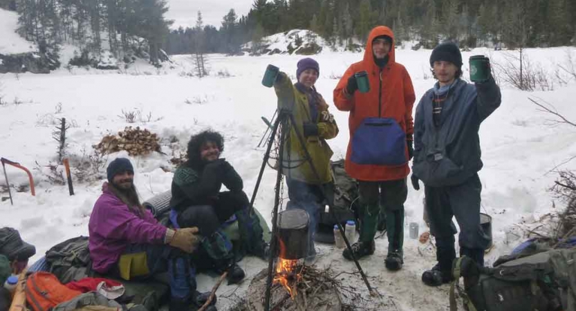 a group of gap year students gathered around a campfire in a snowy landscape raise thermoses in the air
